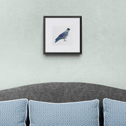 Embroidered bird artwork print of a Greenwich Jackdaw in a black frame, shown as contemporary wall art above a grey sofa in a styled living space.