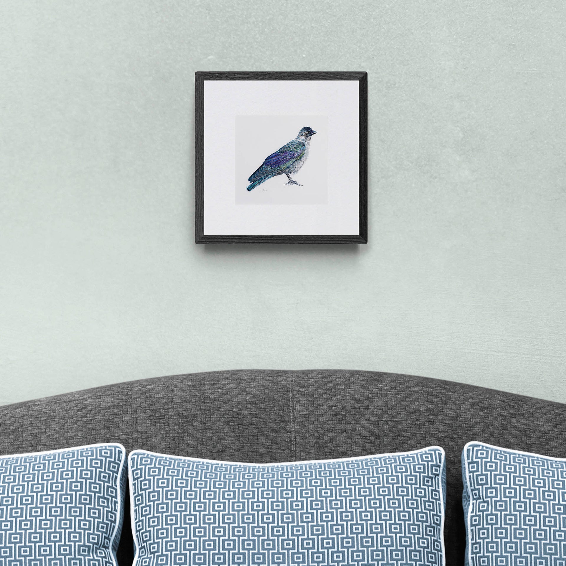 Embroidered bird artwork print of a Greenwich Jackdaw in a black frame, shown as contemporary wall art above a grey sofa in a styled living space.