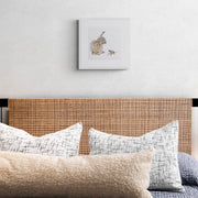 Greenwich Rabbit and Mouse limited edition nature-inspired textile art print by Susannah Weiland, shown in a white frame above a bed with a rattan headboard and layered cream, white and blue-grey textured cushions, the delicate embroidered detail bringing warmth and character to a serene bedroom setting.