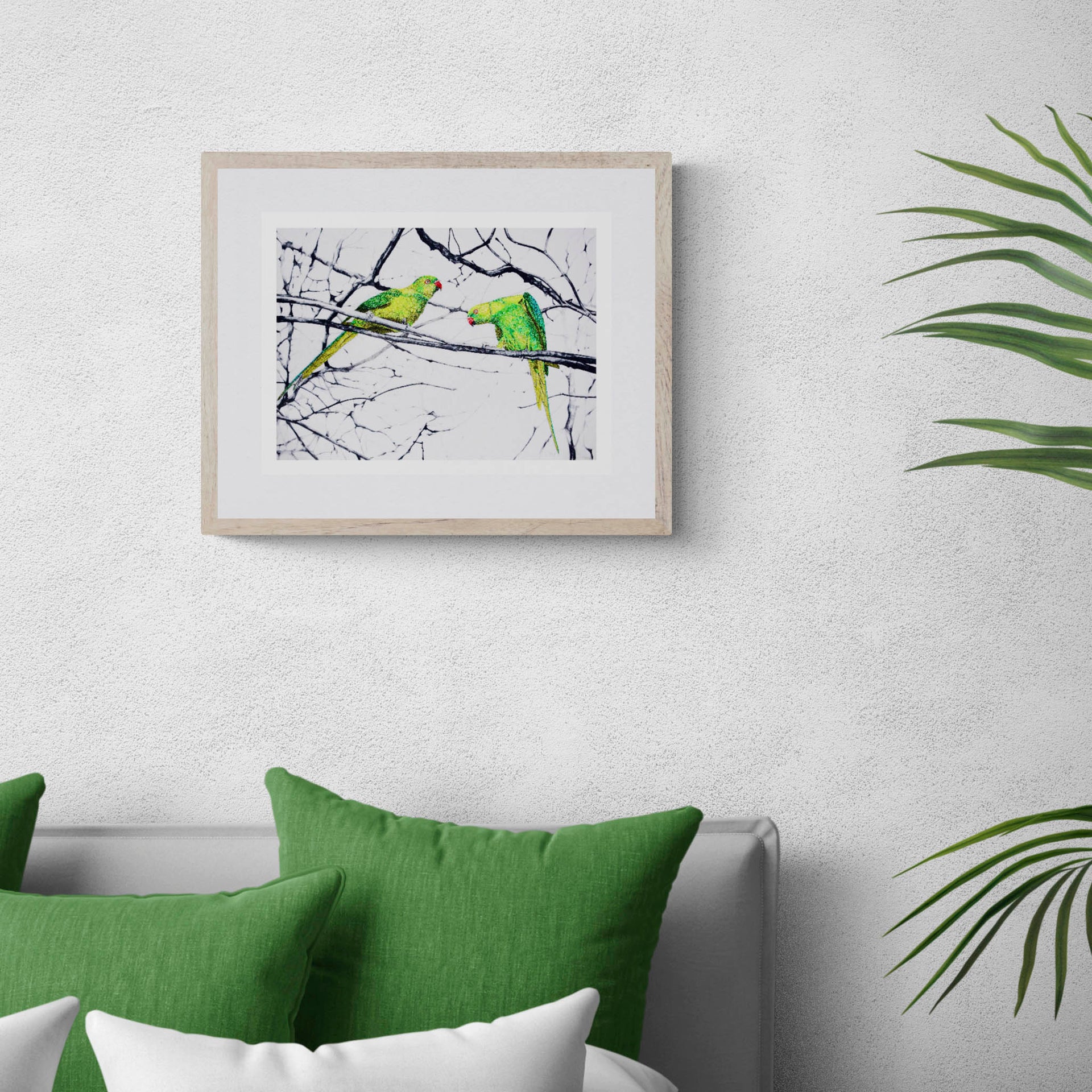 Hyde Park Parakeets limited edition embroidery art print by Susannah Weiland, shown in a natural oak frame with white mount above a sofa with green cushions and tropical foliage, the parakeet subject echoing the surrounding botanics.