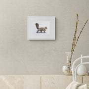 Hyde Squirrel limited edition hand embroidered wall art print by Susannah Weiland, shown in a white frame on a warm taupe wall above a pale wood sideboard with decorative vessels and yellow-flowering branches, the squirrel's warm brown tones echoing the natural materials of the interior.
