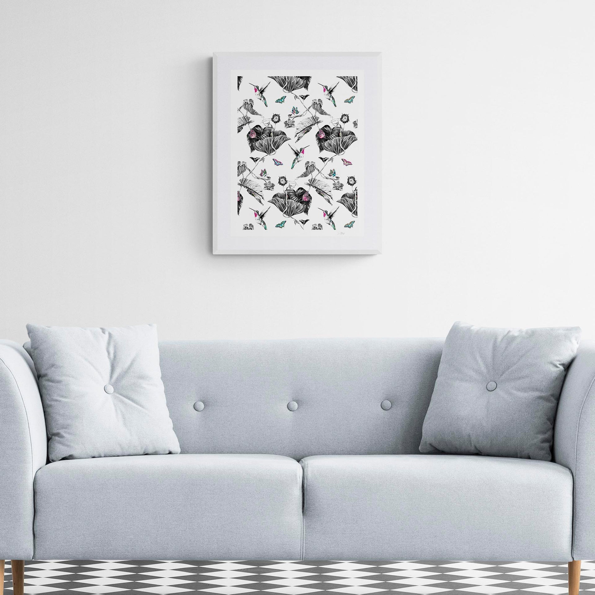 Luxury embroidered artwork by Susannah Weiland in a white frame displayed above a grey button-back sofa in a classic interior with a monochrome diamond-tiled floor. The Pink Hummingbird Limited Edition Wildlife Art Embroidery Print.