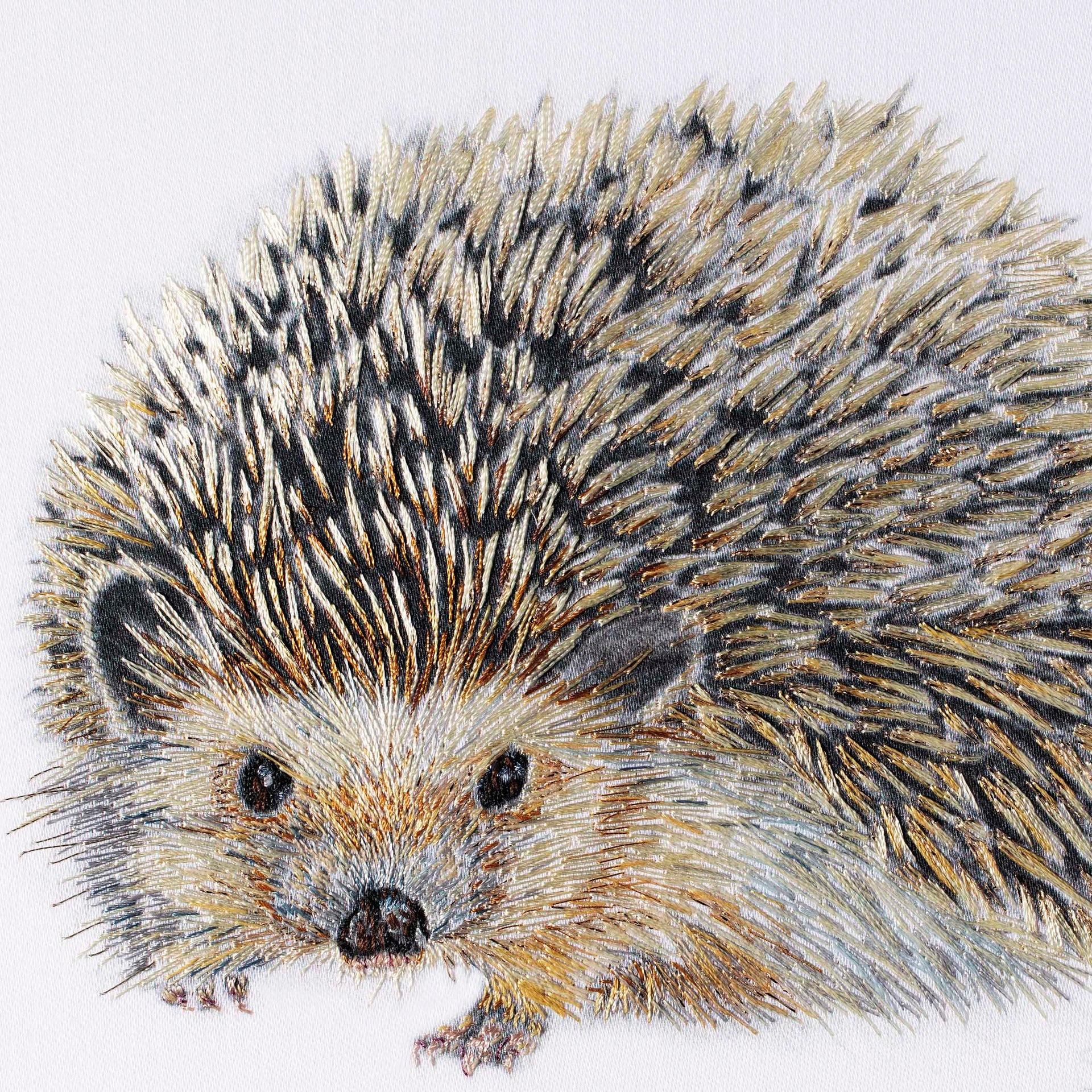 Close-up detail of Regent Hedgehog showing the hand embroidery. Individual stitches in amber, gold, cream and black thread radiate outward across the spines, with metallic threads catching the light. The face is worked in warm golden brown with a dark embroidered nose and bright eyes, the fine stitching around the muzzle capturing the soft fur beneath the spines.