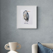 Richmond Riverside Heron limited edition contemporary textile art by Susannah Weiland, shown in a white frame on a dark blue-grey textured wall above a sofa with blue velvet and cream cushions, the heron's palette echoing the moody tones of the surrounding interior.