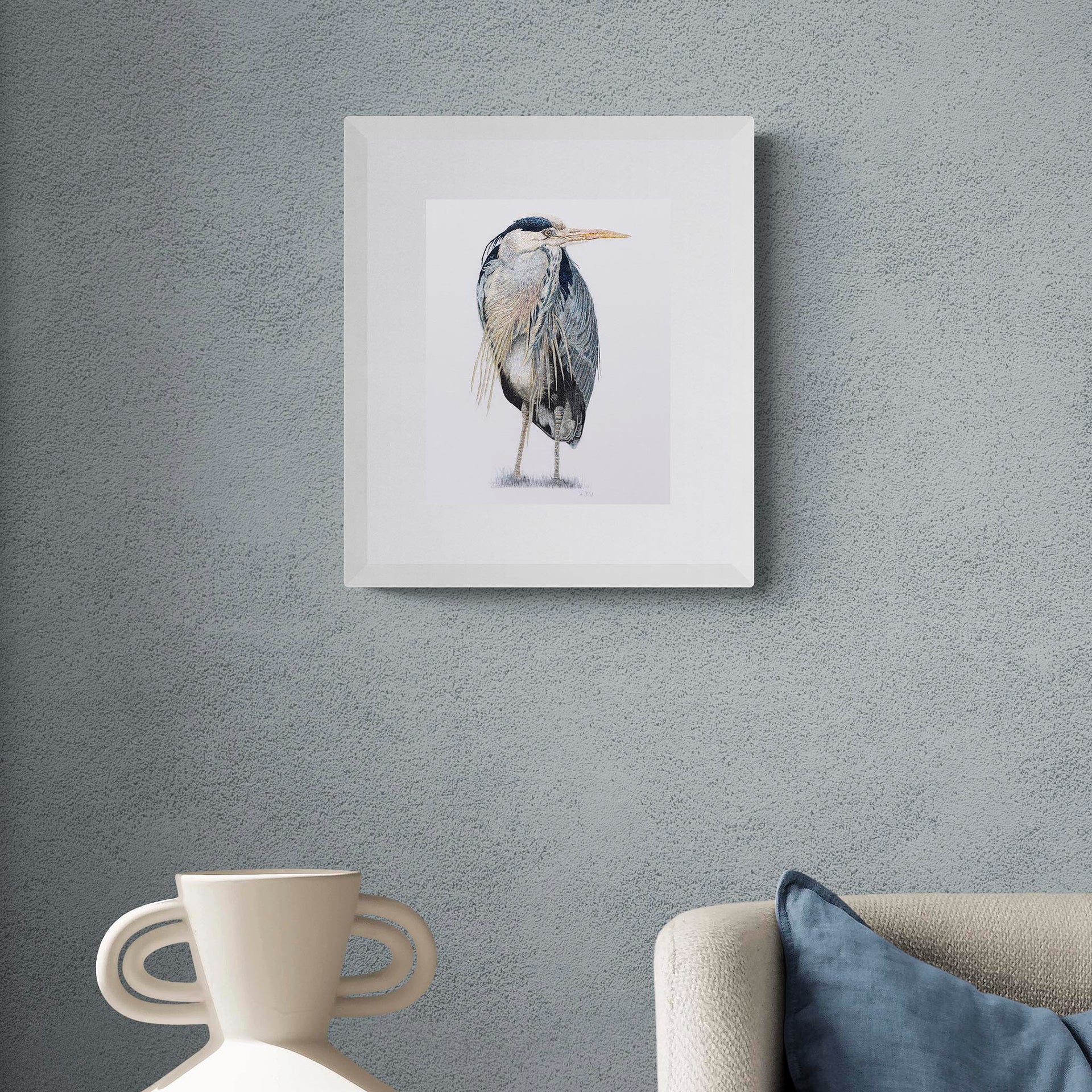 Richmond Riverside Heron limited edition contemporary textile art by Susannah Weiland, shown in a white frame on a dark blue-grey textured wall above a sofa with blue velvet and cream cushions, the heron's palette echoing the moody tones of the surrounding interior.