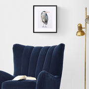 Richmond Riverside Heron limited edition embroidered bird artwork by Susannah Weiland, shown in a black frame with white mount on a white wall beside a navy velvet wingback armchair and brass floor lamp, the combination of bold frame and rich interior creating an elegant, considered display.