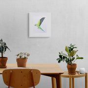 St James Flying Parakeet limited edition hand embroidered wall art print by Susannah Weiland, shown in a white frame on a grey textured wall above a wooden dining table surrounded by terracotta pots and houseplants, the parakeet's tropical energy perfectly at home in a plant-filled, nature-inspired space.