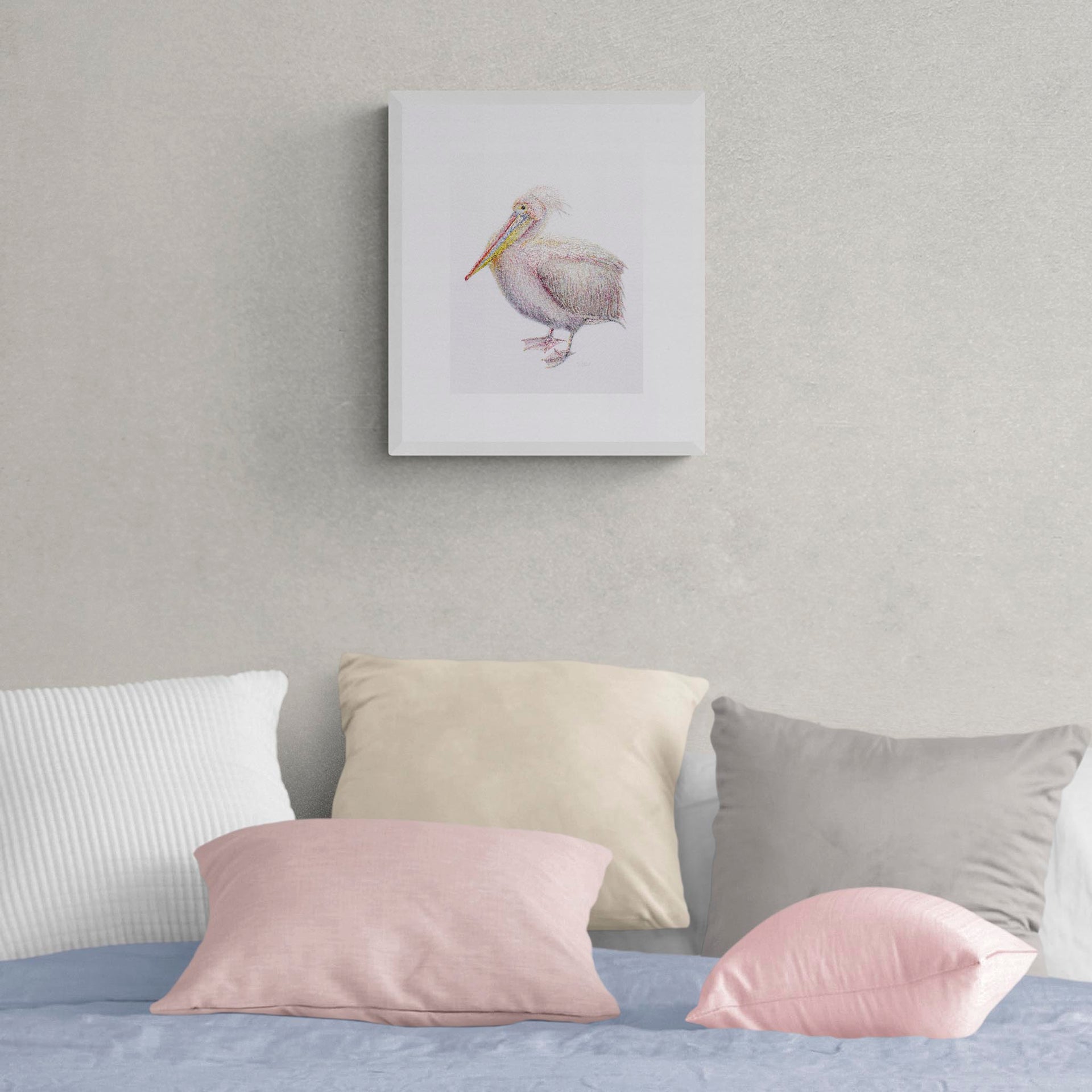 St James Pelican limited edition nature-inspired textile art print by Susannah Weiland, shown in a white frame on a warm grey wall above a bed with pink, cream and grey cushions, the pelican's soft blush tones echoing the gentle bedroom colour palette beautifully.